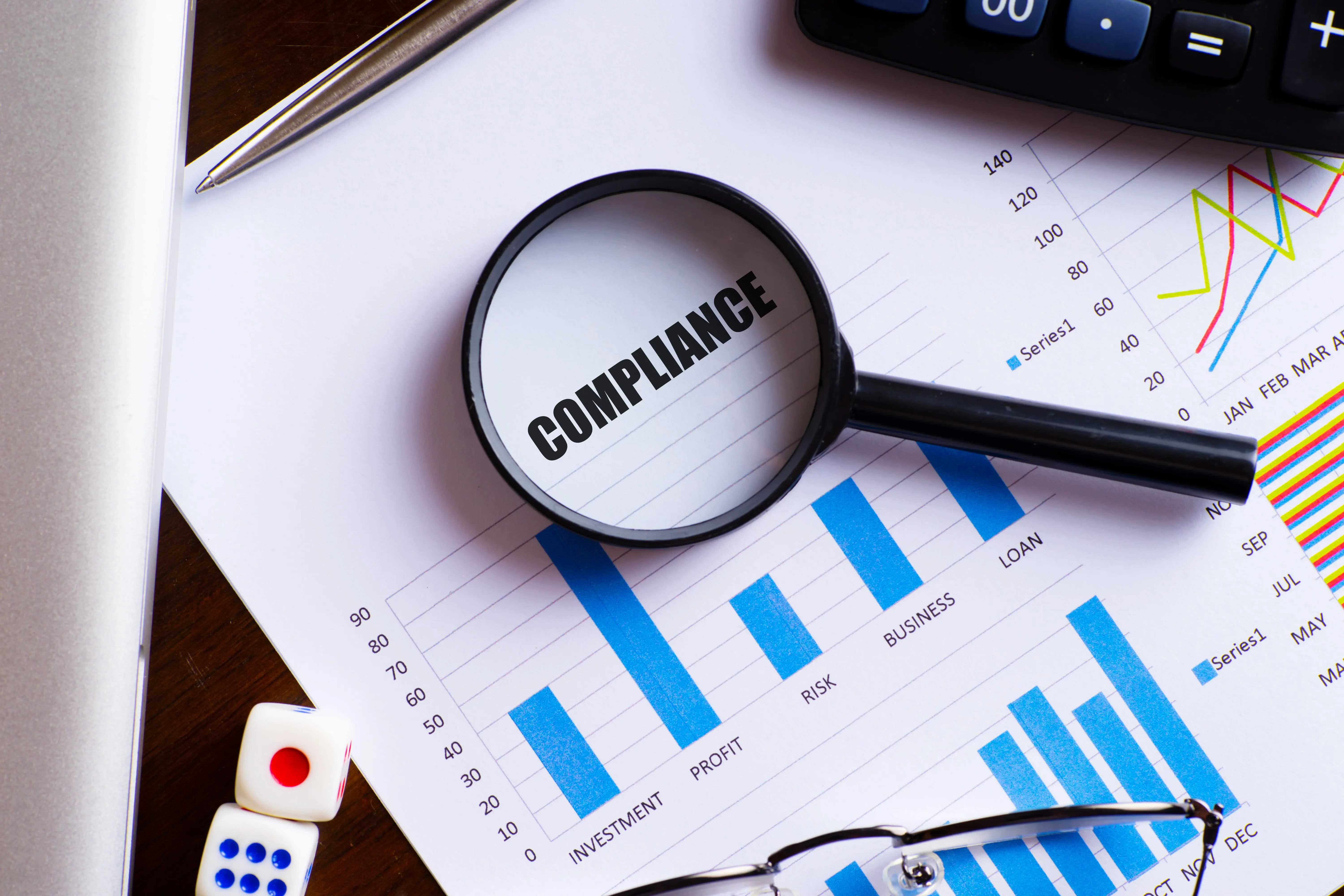 Magnifying glass on "Compliance" text on chart, dice, spectacles, pen, laptop calculator on wooden table - banking, financial services and insurance concept.